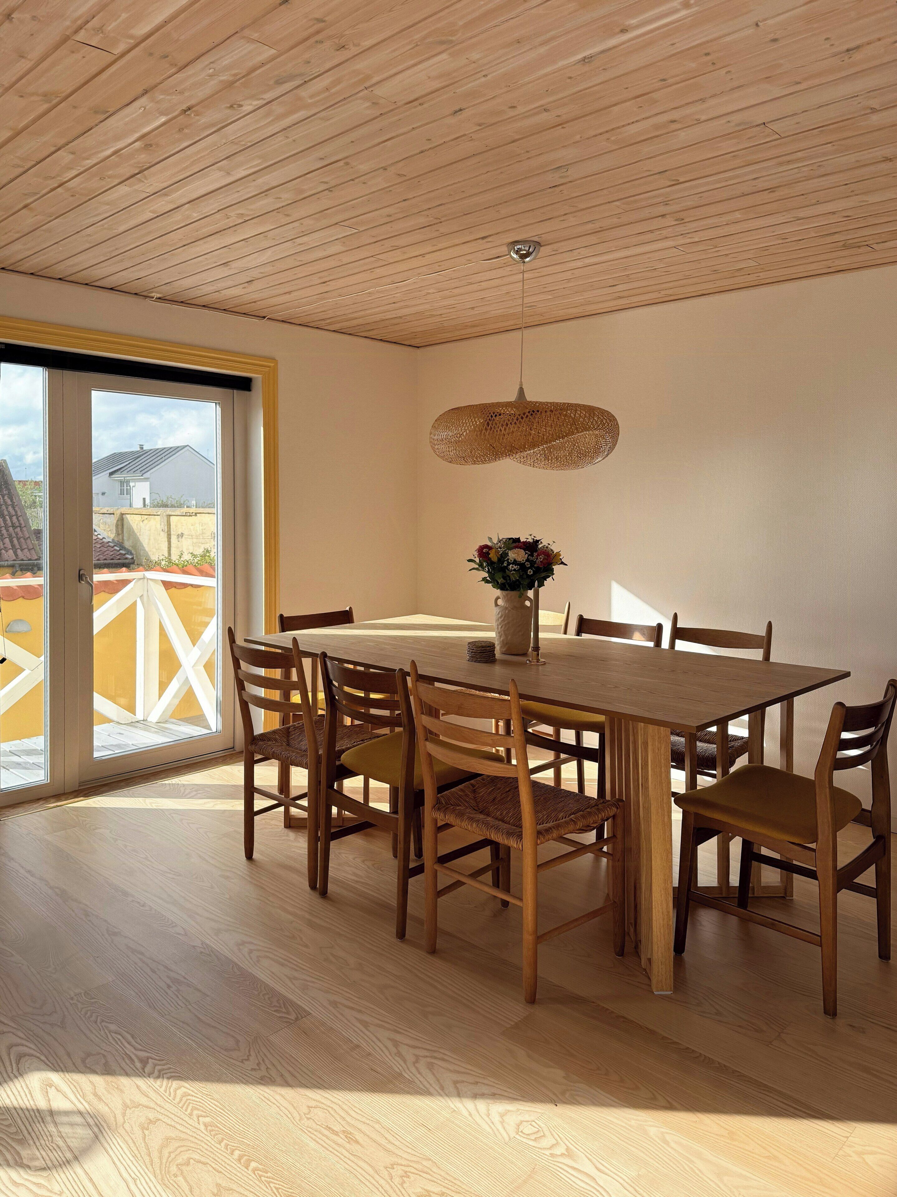 Un comedor con una mesa y sillas de madera, una lámpara colgante y vistas a través de un ventanal.