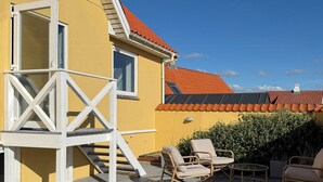 A wooden deck with outdoor furniture, a white staircase, and a yellow building with a red roof.