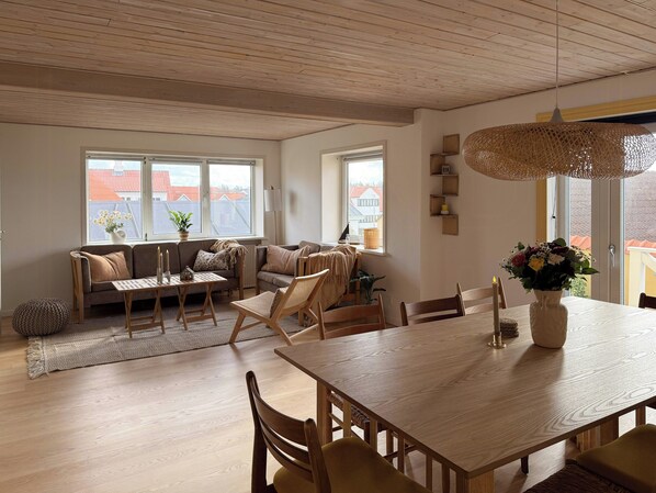 A cozy living room with a wooden ceiling, a sofa, a dining table, and a view of buildings through large windows. - Yellow beach house with sauna and view over Løkken (Løkken)