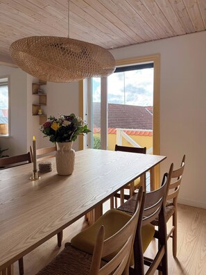 A dining area with a wooden table, chairs, a woven pendant light, and a vase with flowers. - Yellow beach house with sauna and view over Løkken (Løkken)