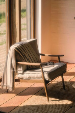A modern chair with a cushion and a blanket, positioned on a wooden floor near a window. - Eco-friendly cabin in the Norman countryside (Gathemo)