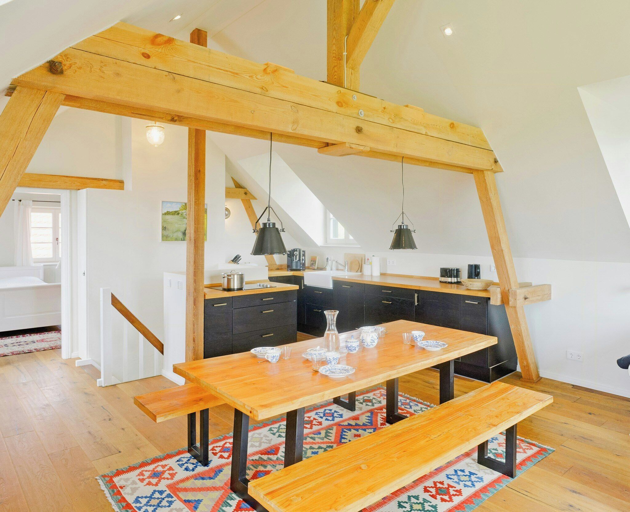 A loft-style kitchen with exposed wooden beams, a wooden dining table with benches, and a hanging pendant light.