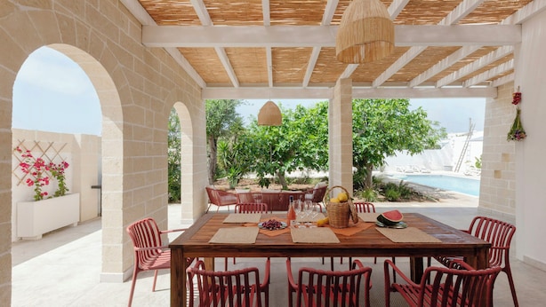 Area esterna con zona pranzo, tavolo in legno e sedie rosse, circondata da muri in pietra e vista su una piscina.