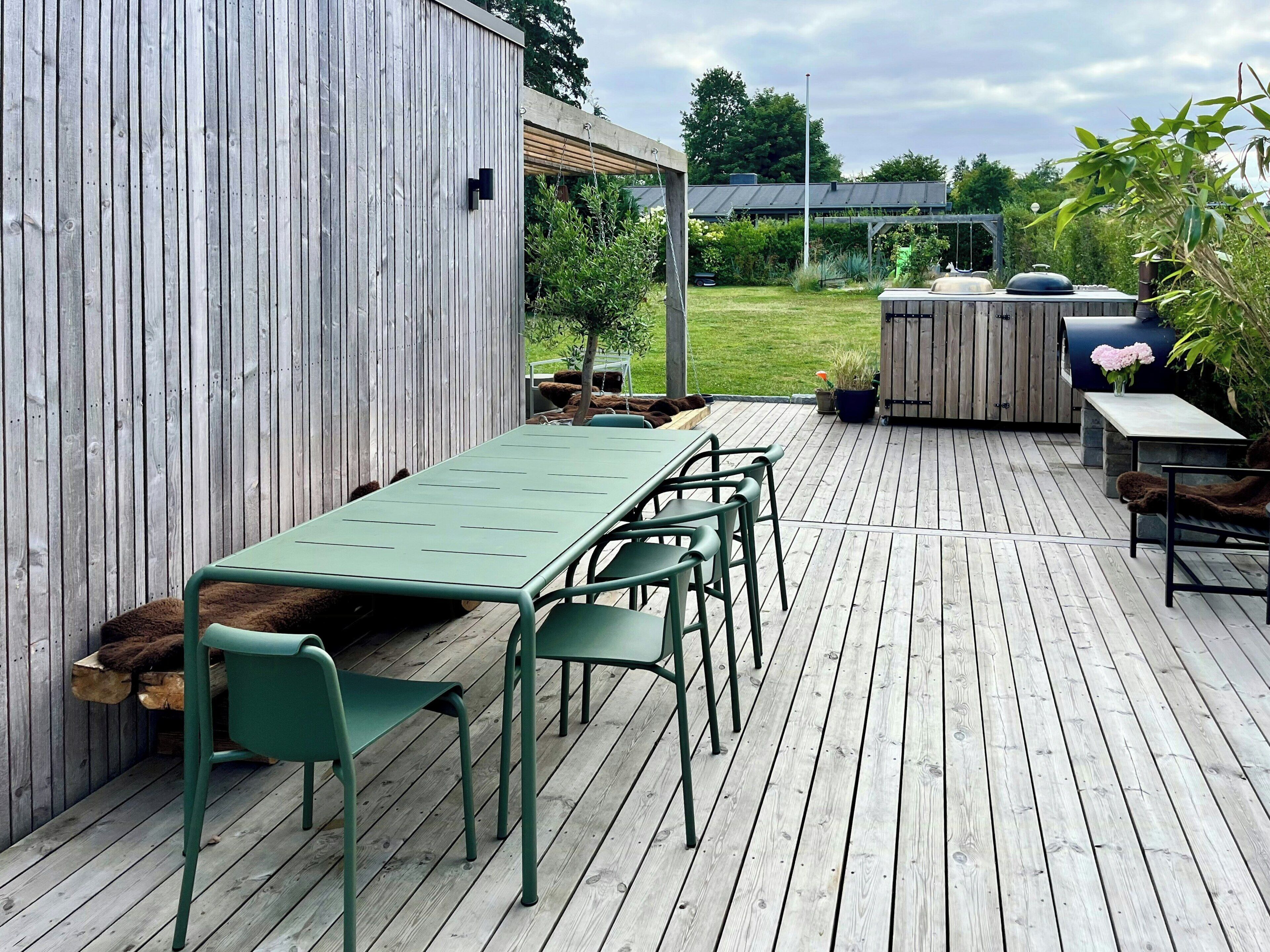 Una terraza de madera con una mesa, sillas, una parrilla y un jardín al fondo.