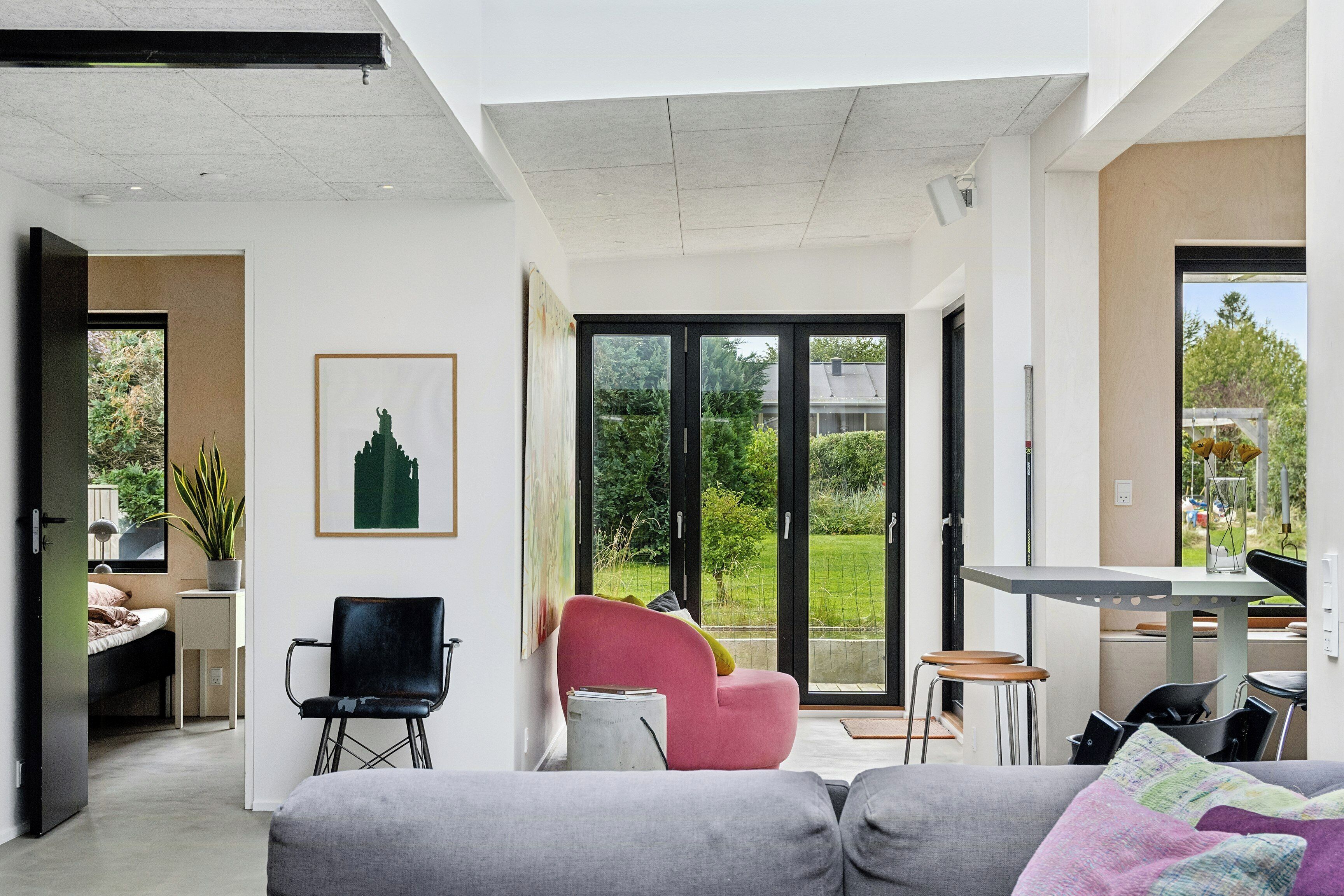 A modern living room with a grey sofa, a pink chair, a dining table, and a sliding glass door leading to a garden.