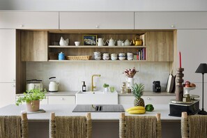 A modern kitchen with white cabinetry, a wooden shelf with kitchenware, a white sink, and a dining area with wicker chairs. - New villa with pool in Sainte-Marine (Combrit)