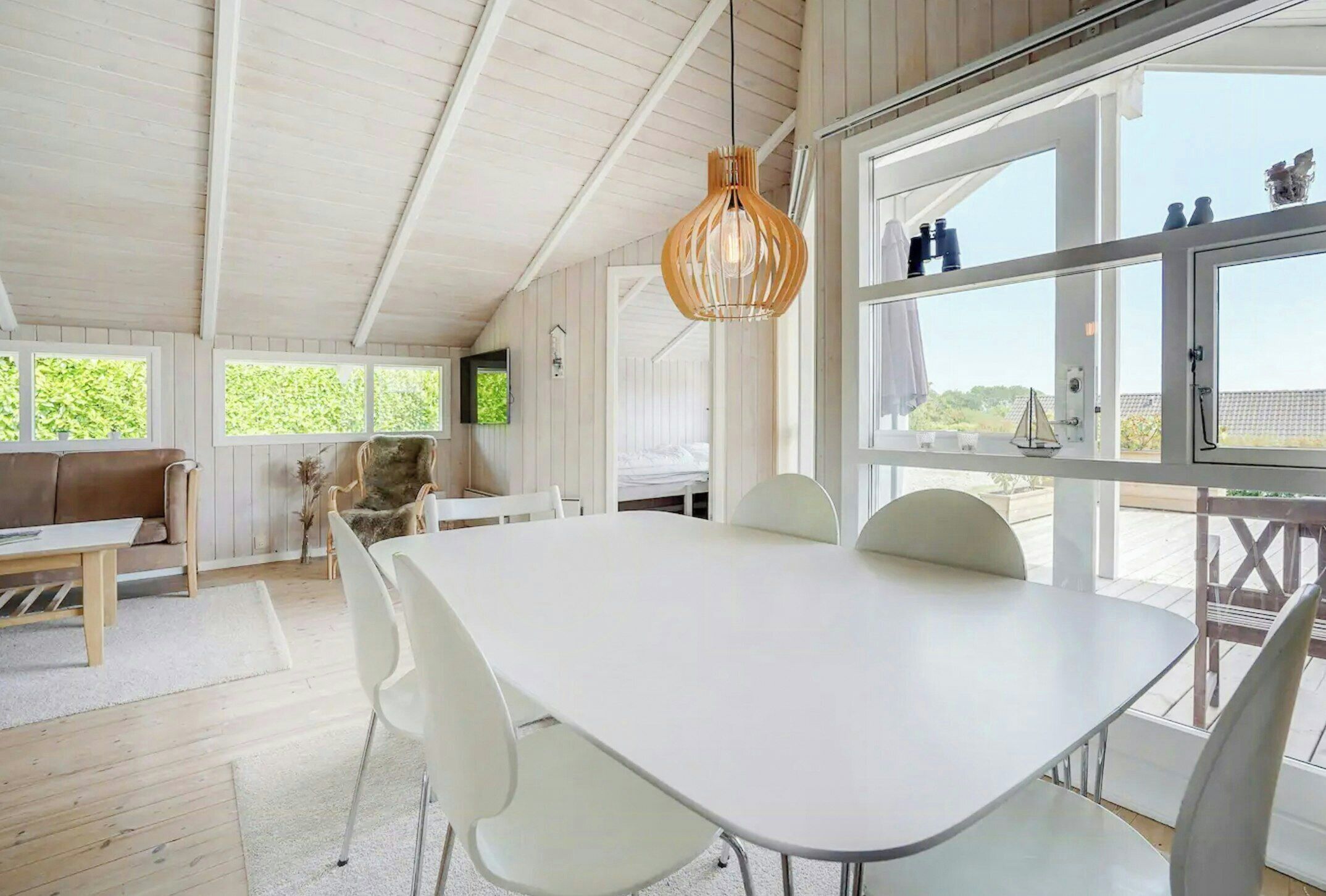 A spacious room with a large dining table, white chairs, a hanging light fixture, and large windows offering a view of the outdoors.