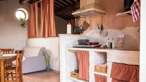 A kitchen with a dining table, chairs, a hanging pot rack, and a window with curtains. - Panoramic refuge on Monte Pelato (Gualdo Cattaneo)
