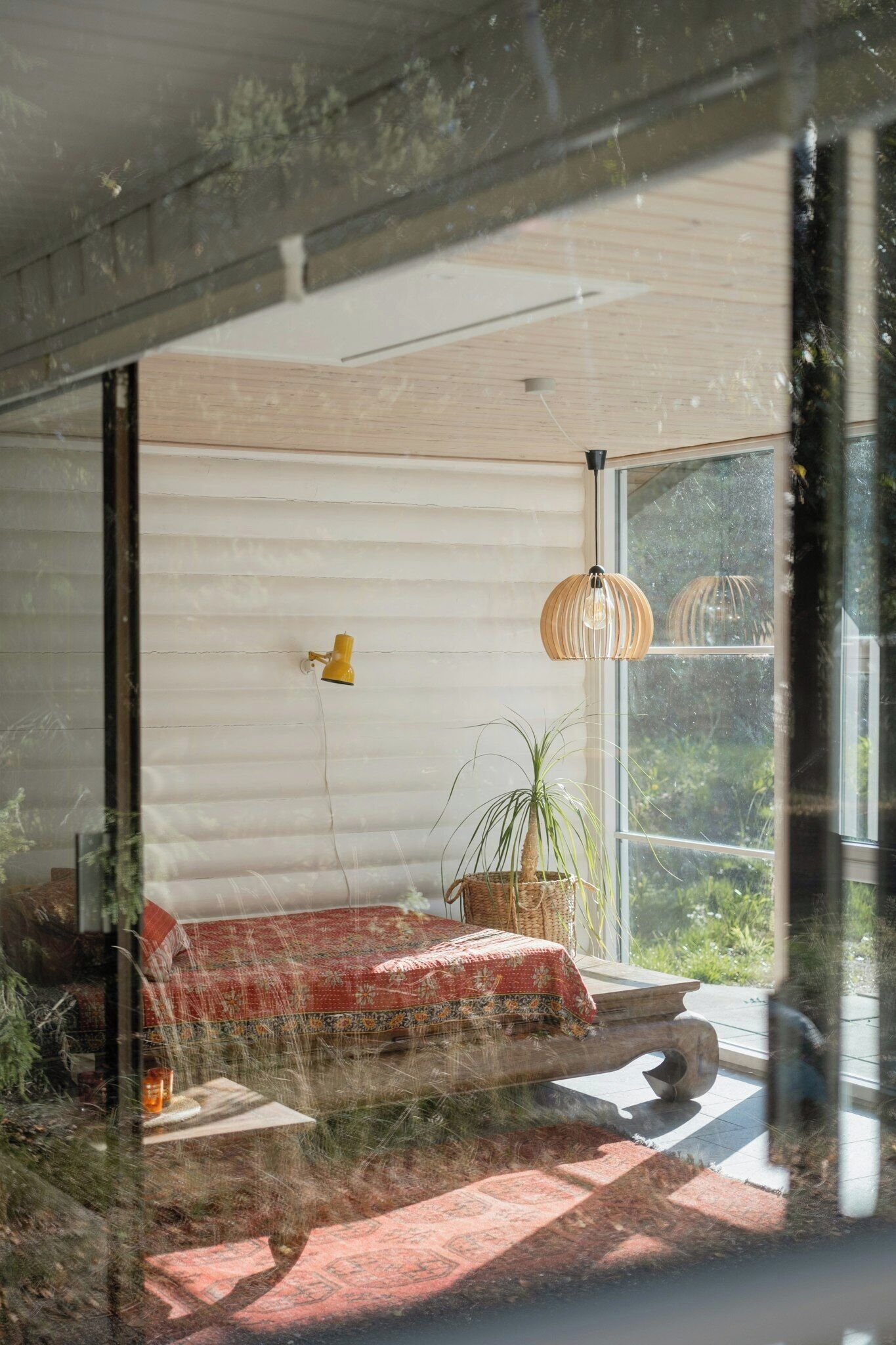 Una habitación con cama, planta en maceta, lámpara colgante y ventana con vista a follaje.