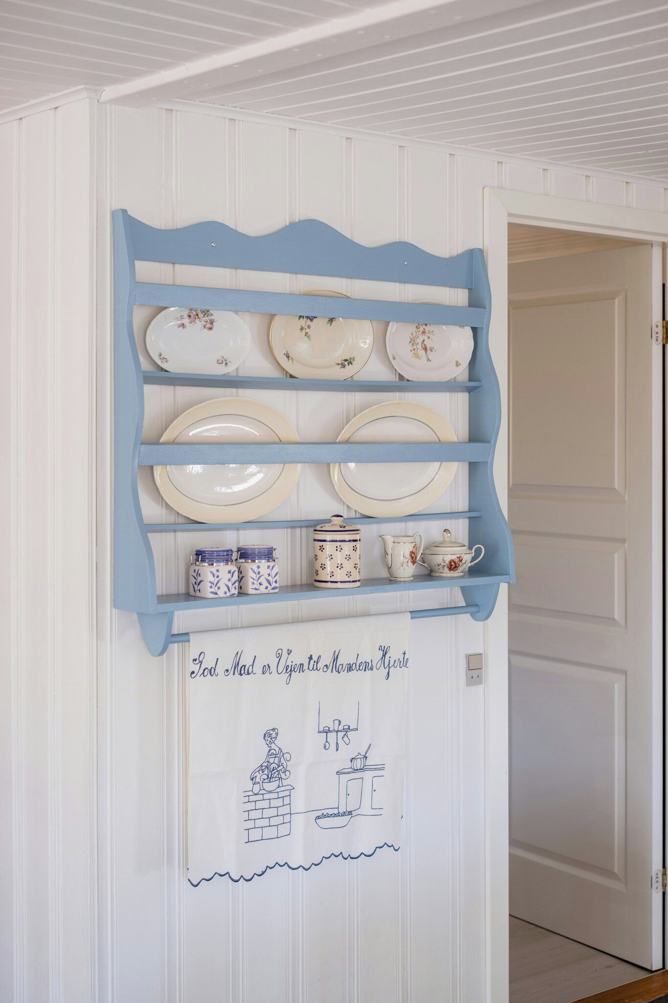 A kitchen area with a blue shelf holding plates and jars, a white door, and a wall with a handwritten note.