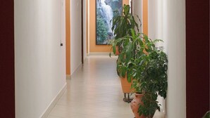 Um corredor com piso de azulejos, uma grande planta em vaso e um quadro na parede.