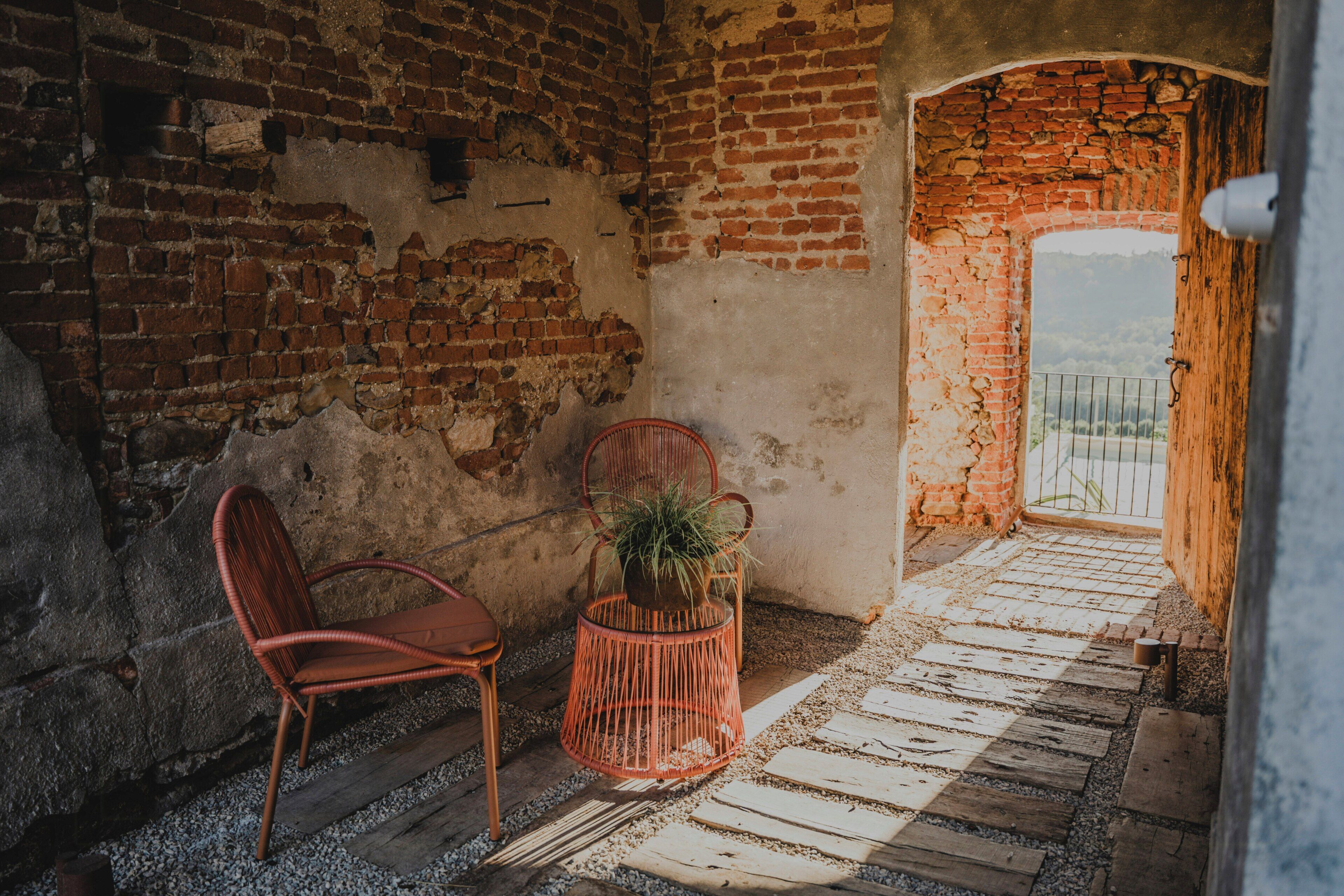 Un espacio interior con pared de ladrillo, una silla roja, una silla de mimbre y una planta en maceta.