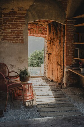 An open doorway leading to a balcony with a view, a wicker chair, a potted plant, and a rustic wooden shelf. - Farmhouse among hazelnut groves in the Langhe (Cherasco)