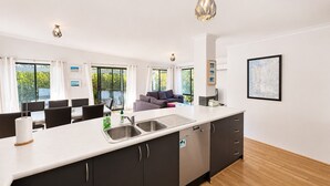 A modern kitchen with a large island, a dining area with a table and chairs, a sofa, and a view of greenery outside. - Busso Escape – The Perfect South West Getaway (Broadwater)