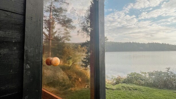 A wooden cabin with a glass door, a wooden floor, and a view of a lake and trees. - Mini house with sandy beach and sauna (Olofstorp)