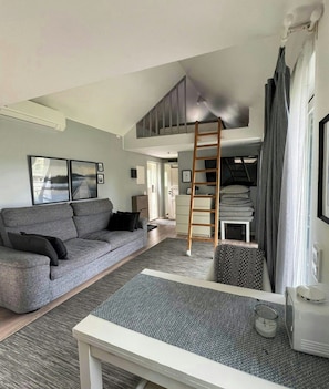 Ein modernes Wohnzimmer mit grauem Sofa, weißem Couchtisch, einer Leiter und einem Schlafplatz mit Stauraum.