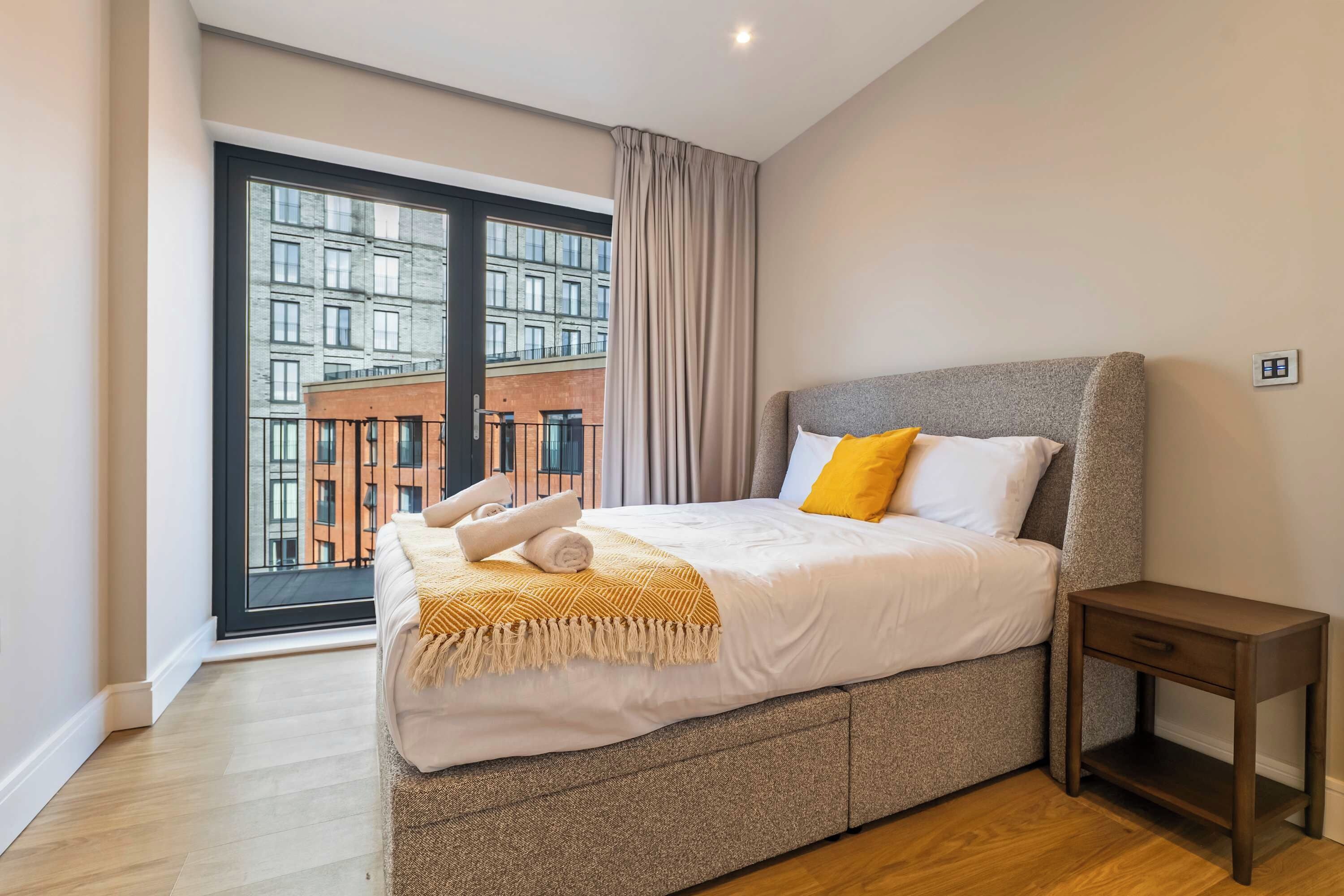 A modern bedroom with a large window, a bed with white bedding and a yellow pillow, a grey headboard, and a wooden nightstand.