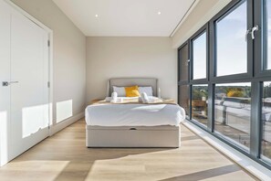 A modern bedroom with a large bed, a nightstand, and a view through floor-to-ceiling windows. - Spacious 3BR Near Bullring | StayCozy (Birmingham)