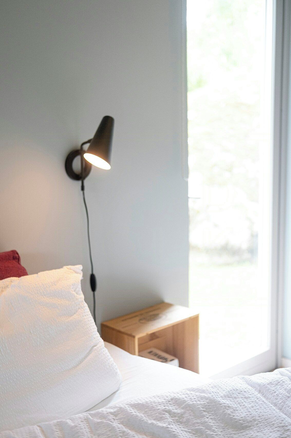 A bed with white bedding, a bedside table with a lamp, and a window with a view of greenery.