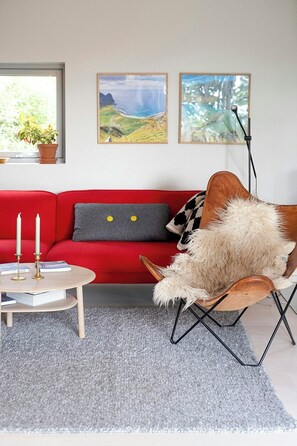 A red sofa with a grey pillow, a wooden chair with a fur throw, a small round table with books and candles, and two framed landscape paintings on the wall. - Modern flat in Kabelv åg, Lofoten (Nordland)