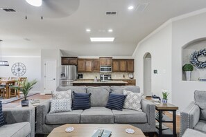 A living room with a grey sofa, a coffee table, and a dining area with a wooden table and chairs. - Bogey`s House - 3BR/2BA Brand New Listing (Lubbock)