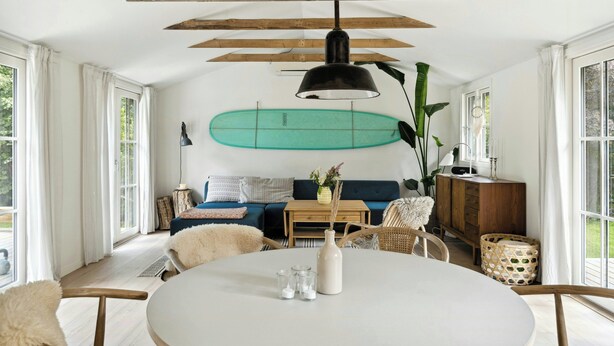 Ein Zimmer mit einem runden Tisch, einer Couch, einer Holzbalkendecke und einem Surfbrett an der Wand.