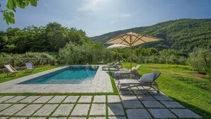 Una piscina con sillas de descanso y una sombrilla, rodeada de vegetación y colinas.