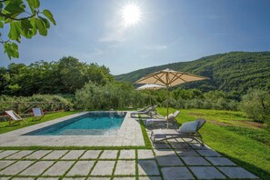 Ein Pool mit Liegestühlen und Sonnenschirm, umgeben von viel Grün und Hügeln.