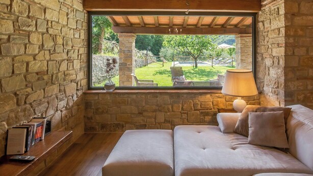 Una habitación con paredes de piedra, un banco de madera, un sofá y una ventana con vistas a un jardín.
