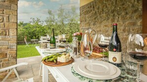 Un espacio de comedor al aire libre con un muro de piedra, una mesa puesta con platos, una botella de vino Cuna, una jarrón y una copa de vino tinto.