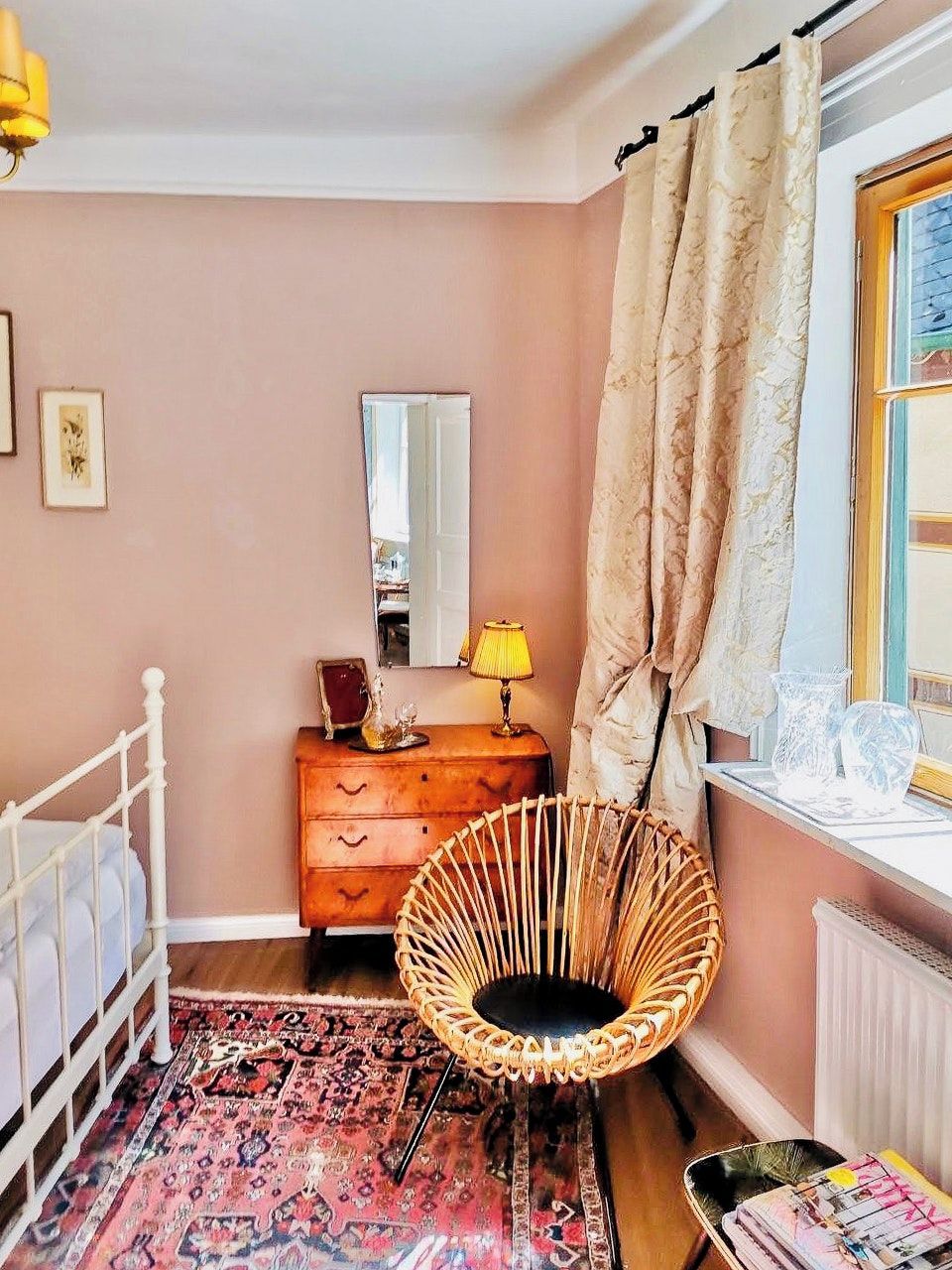 A bedroom with a white bed, a wooden dresser, a wicker chair, a mirror, and a window with curtains.