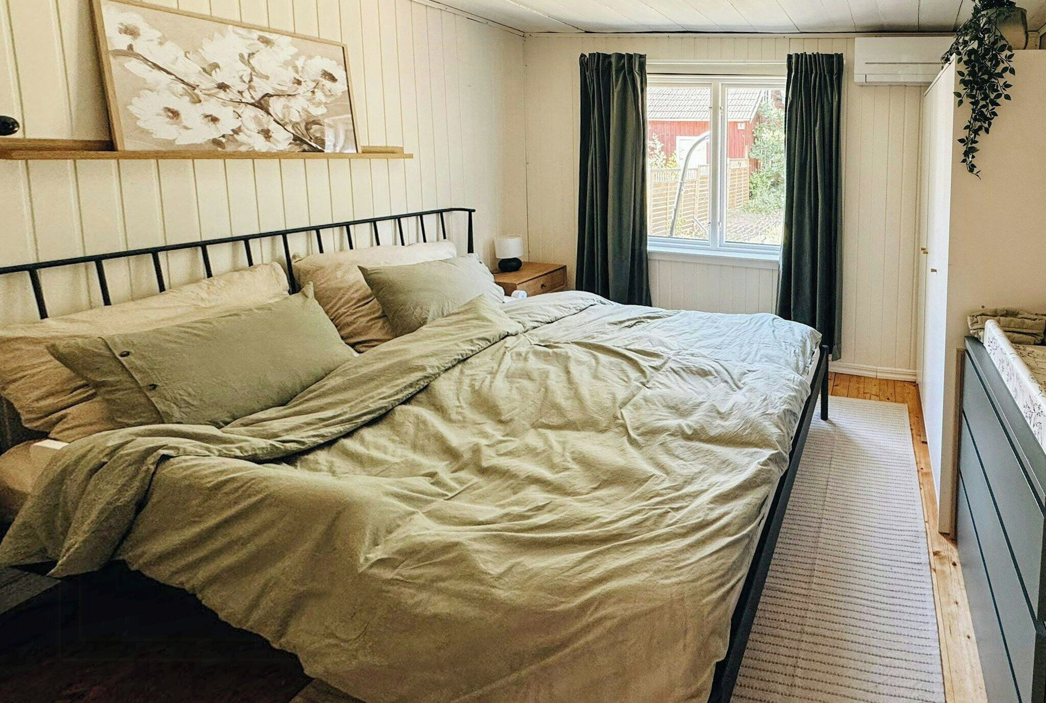 A bedroom with a bed, a nightstand, a window with curtains, and a wall-mounted plant.