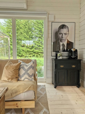 A room with a wooden floor, a sofa with patterned cushions, a coffee maker on a dark cabinet, and a poster on the wall.