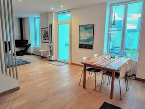 Ein modernes Wohnzimmer mit Holzboden, einem Esstisch mit Stühlen, einer schwarzen Couch, einem Fernseher und einem großen Fenster mit Aussicht.