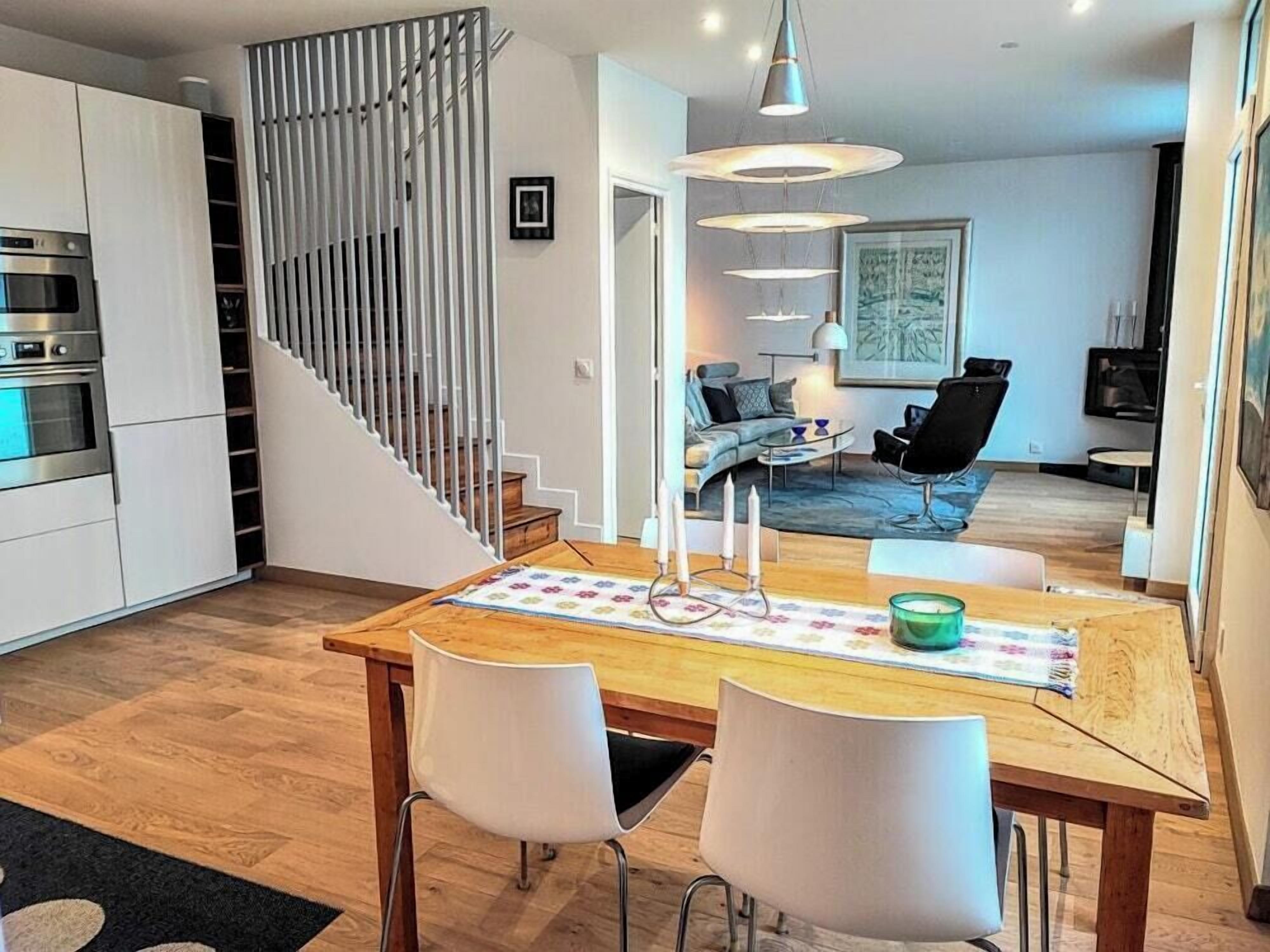 Une cuisine moderne dotée d’appareils électroménagers intégrés, un coin repas avec une table en bois et des chaises blanches, et un salon avec un canapé et un téléviseur.