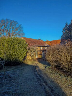 Ein ländliches Gebäude mit rotem Ziegeldach, umgeben von dichter Vegetation und einem Feldweg, der zum Eingang führt.