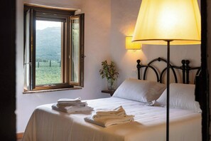 Ein Schlafzimmer mit Bett, Lampe, Fenster mit Aussicht und Nachttisch mit Vase.