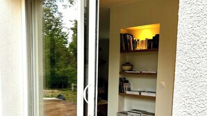 Ein Zimmer mit einer Schiebetür aus Glas, die auf eine Holzterrasse führt, einem Bücherregal mit Büchern und einer Wand mit strukturierter Oberfläche.