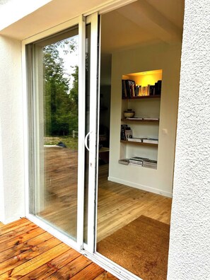 A room with a sliding glass door leading to a wooden deck, a bookshelf with books, and a white textured wall. - Charming house in Lyons-la-Forêt (Lyons-la-Forêt)