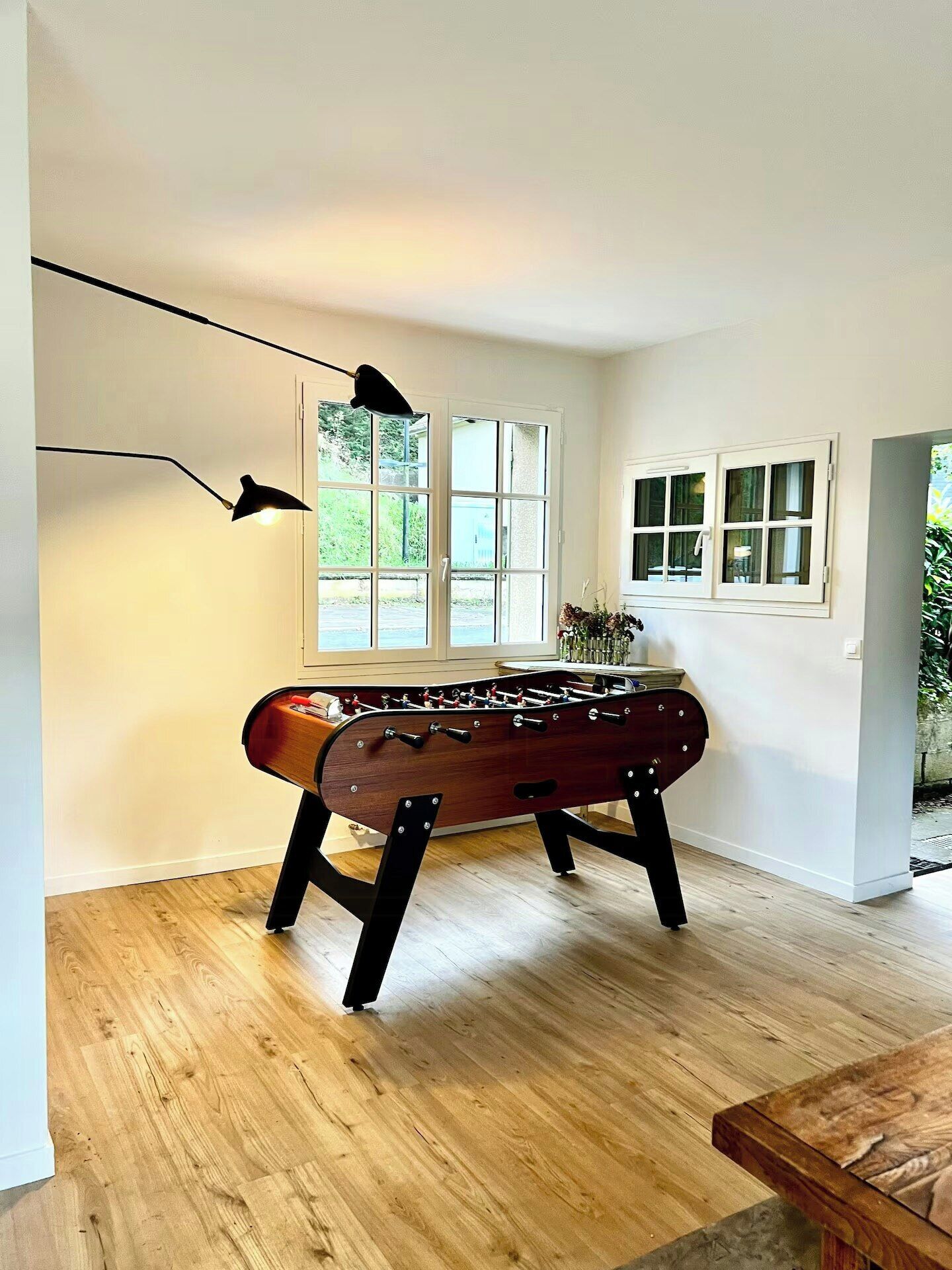 Ein Zimmer mit Holzboden, einem Kicker, einer Lampe und Fenstern mit Blick auf Grünflächen.