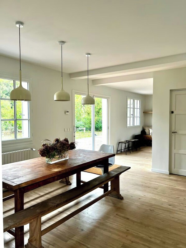 A spacious room with a wooden dining table, hanging pendant lights, and large windows offering a view of greenery outside. - Charming house in Lyons-la-Forêt (Lyons-la-Forêt)
