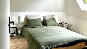 Ein Schlafzimmer mit schrägen Wänden, einem Oberlicht, einem Bett mit grüner Bettwäsche, einem Sessel und einem kleinen Tisch.
