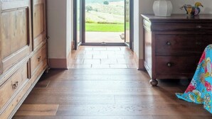 A hallway with a wooden floor, an open doorway leading to a view of greenery, a wooden cabinet, a lamp, and a framed picture on the wall. - Traditional Tuscan villa on hill with pool (Magliano in Toscana)