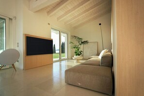 A modern living room with a flat-screen TV, a sofa, a potted plant, and a large window. - Villa with infinity pool overlooking Lake Garda (Bardolino)