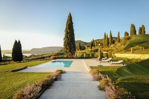 A pool with a paved pathway leading to lounge chairs, surrounded by manicured lawns and tall trees. - Villa with infinity pool overlooking Lake Garda (Bardolino)