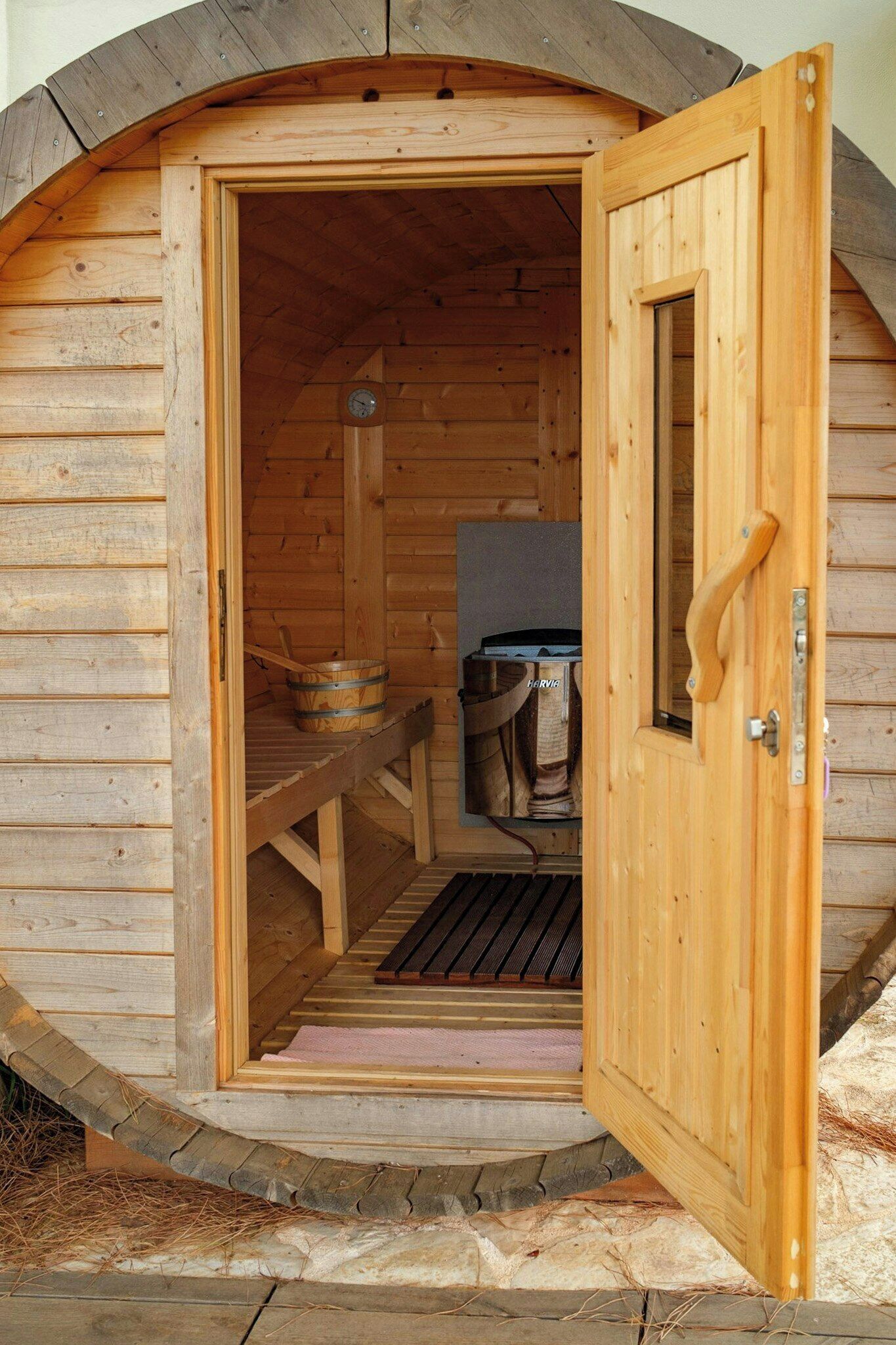 A wooden sauna with a door open, a bench inside, and a bucket outside.