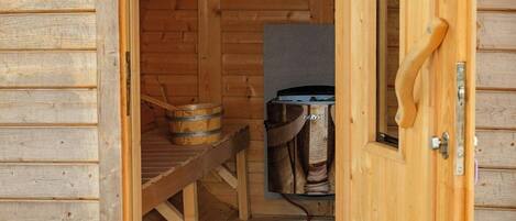 A wooden sauna with a door open, a bench inside, and a bucket outside.