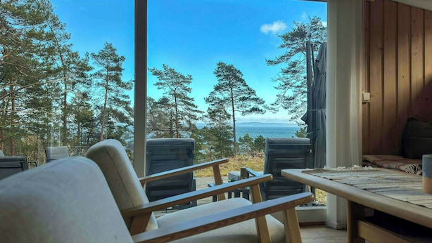 Una habitación con un ventanal grande que da a un bosque y una masa de agua. Hay dos sillas y una mesa con una manta.