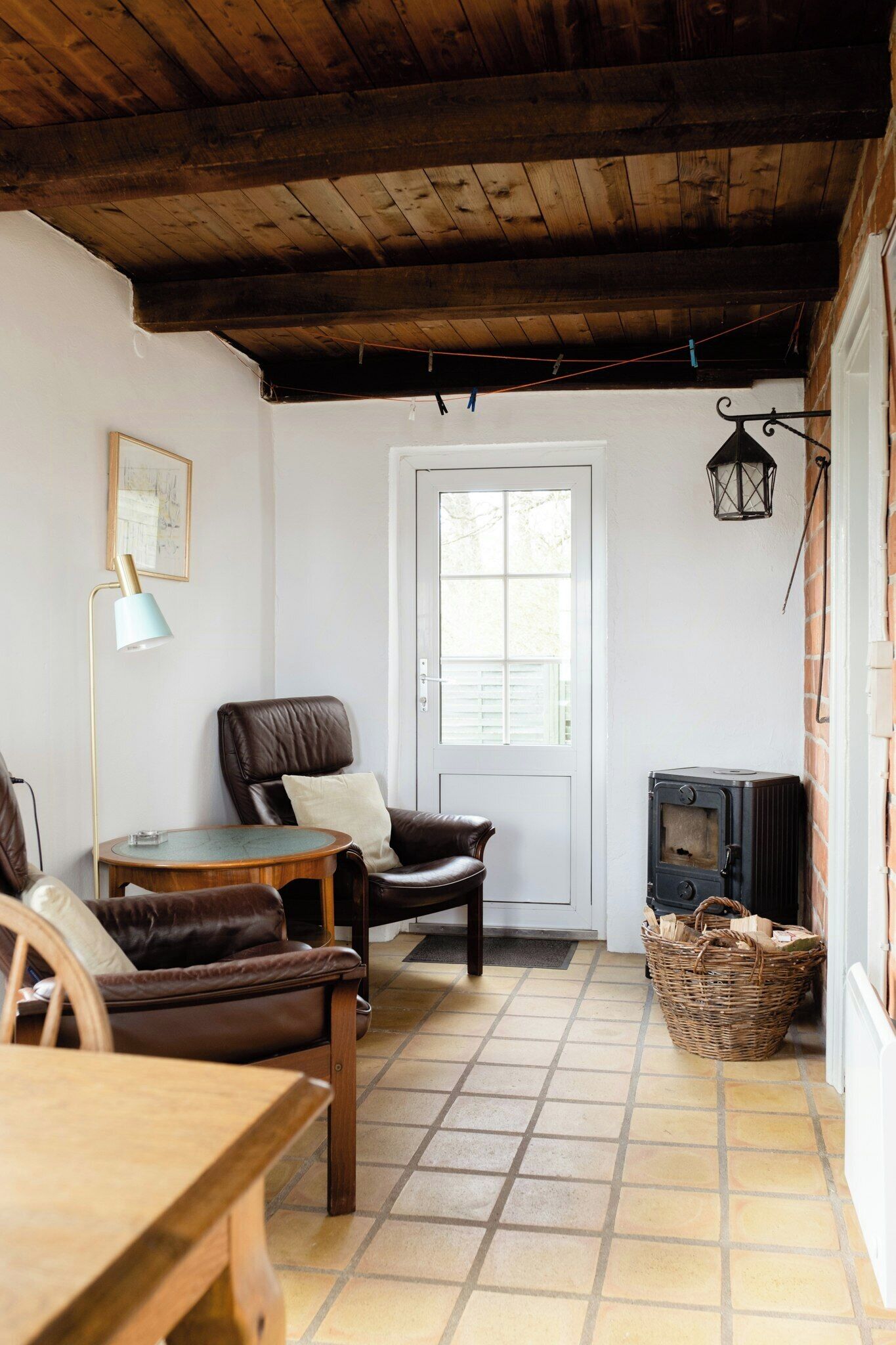 A cozy room with a wooden ceiling, a fireplace, a door, a chair, a table, and a lamp.