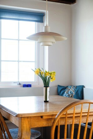 Ein Esstisch mit Stühlen, eine hängende Pendelleuchte, eine Vase mit Blumen und ein Fenster mit blauen Vorhängen.